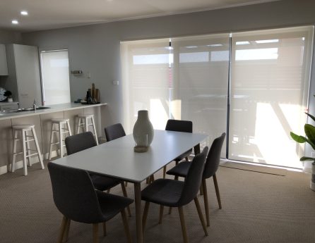 Light roller blinds designed by Australian Plantation Shutters installed beside a dining table and chairs.